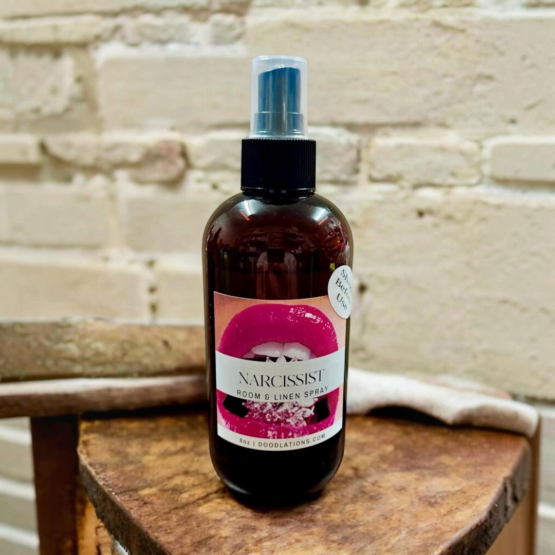 Doodlations Narcissist room and linen spray in amber bottle with bold pink lips label. Fruity-floral scent.