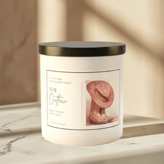 Doodlations The Outlaw hand-poured candle in white jar with black lid, featuring cowboy hat and boots graphic. Masculine scent.