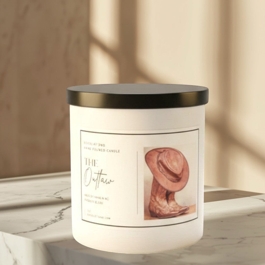Doodlations The Outlaw hand-poured candle in white jar with black lid, featuring cowboy hat and boots graphic. Masculine scent.