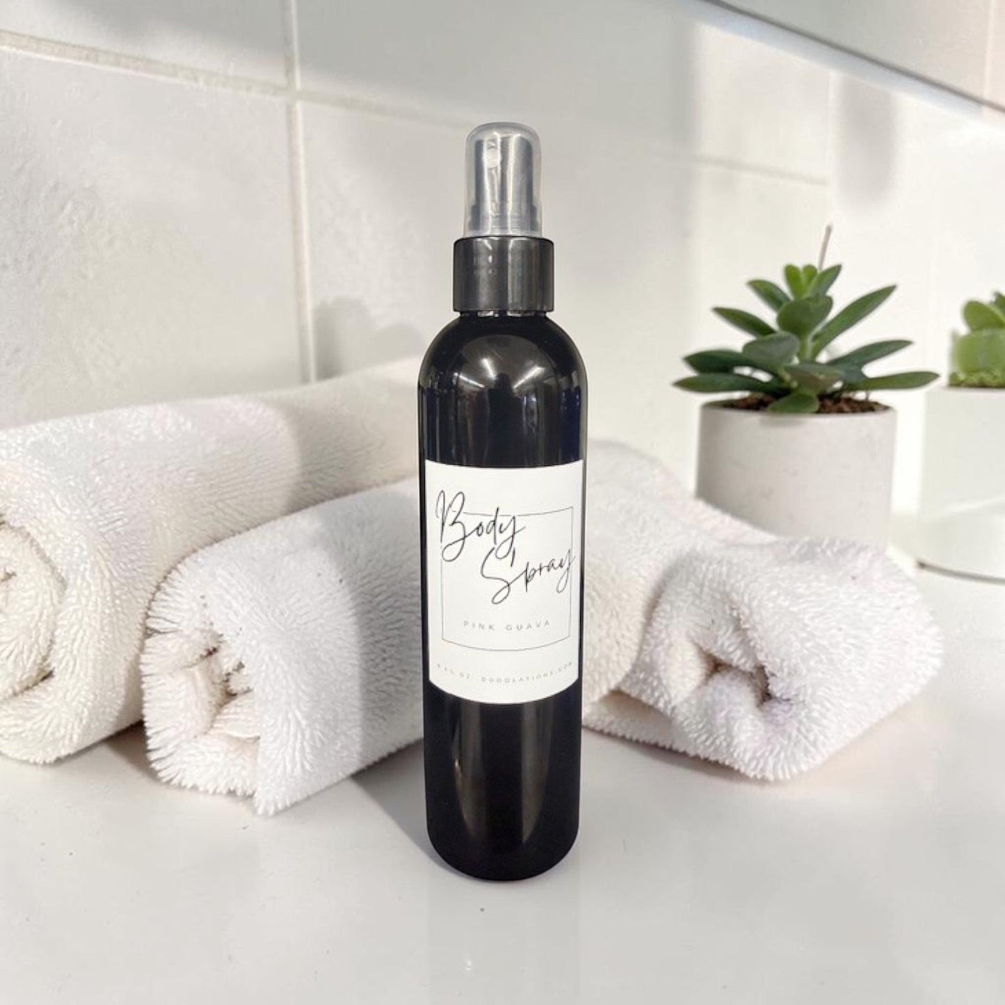 Doodlations Coffee Bar & Boutique Pink Guava Moisturizing Body Spray in a black bottle with a white label, next to rolled white towels and a small potted succulent.