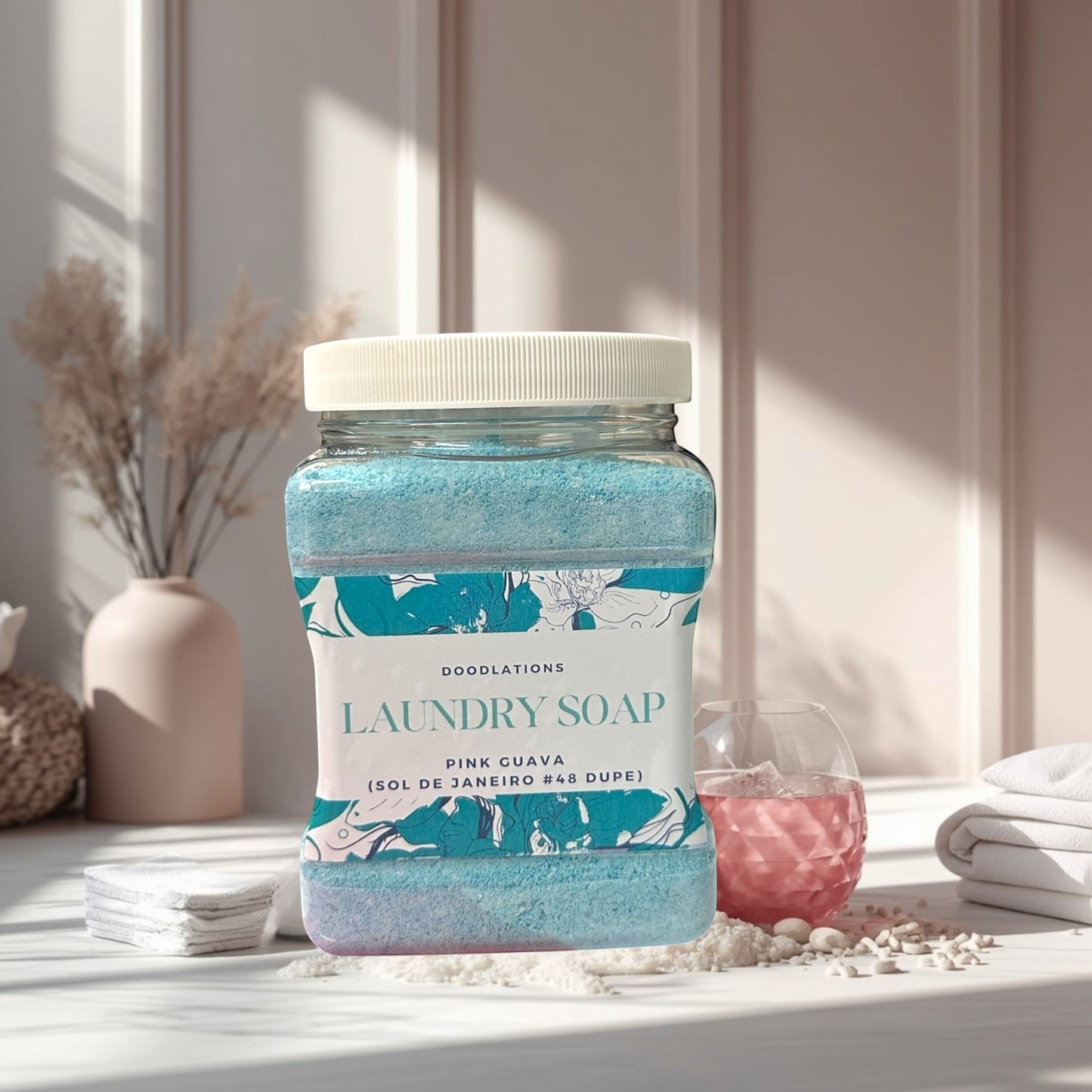 Doodlations Luxury Laundry Soap in Pink Guava scent, a blue granular powder in a clear jar with a white lid. Stack of white towels in the background.