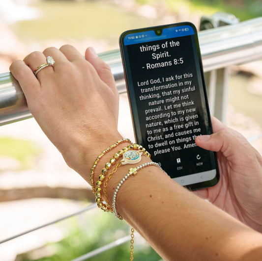 Doodlations Coffee Bar & Boutique Survivor Series Bracelet with cross charm and NFC chip, gold and silver beads, on a wrist holding a phone displaying a Bible verse.