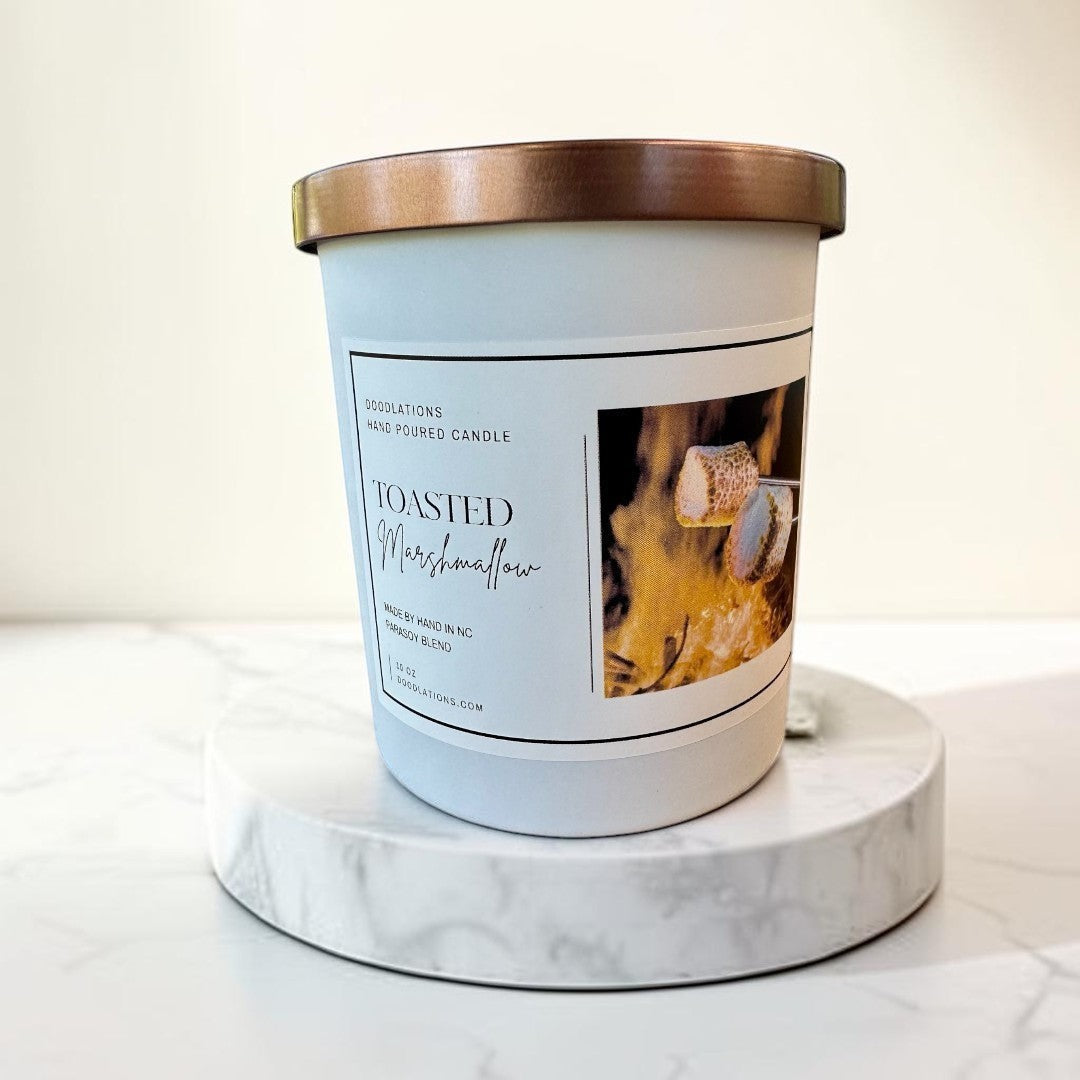 Toasted Marshmallow hand-poured candle with copper lid, set by a campfire with marshmallows.