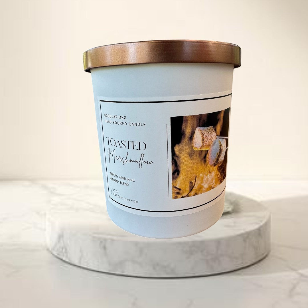 Toasted Marshmallow hand-poured candle with copper lid, set by a campfire with marshmallows.