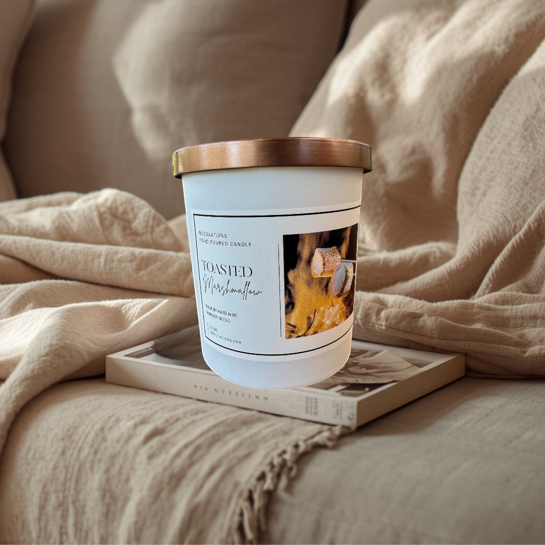 Doodlations Toasted Marshmallow hand-poured candle with a copper lid, set on a wooden table near a campfire.