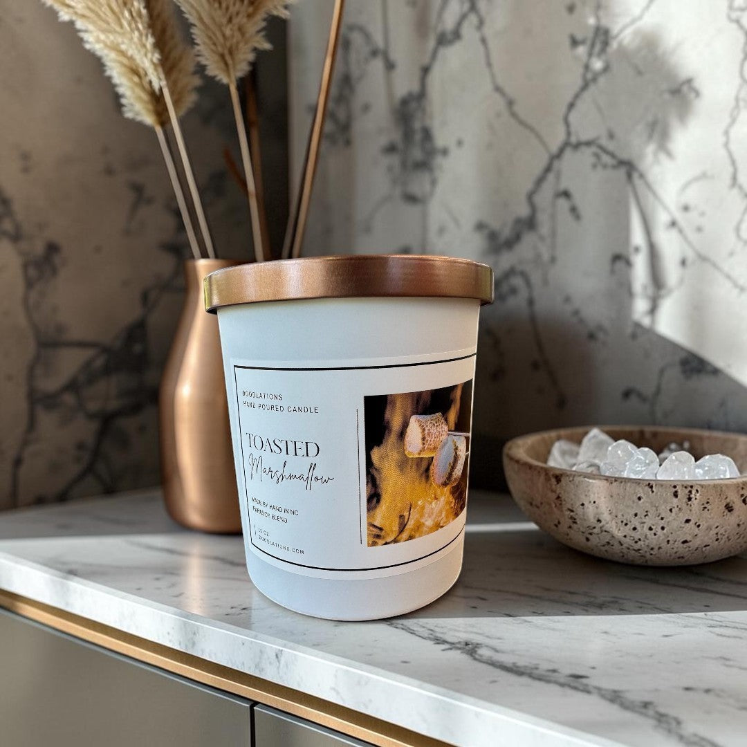 Doodlations Toasted Marshmallow hand-poured candle with copper lid, set by a campfire with marshmallows.