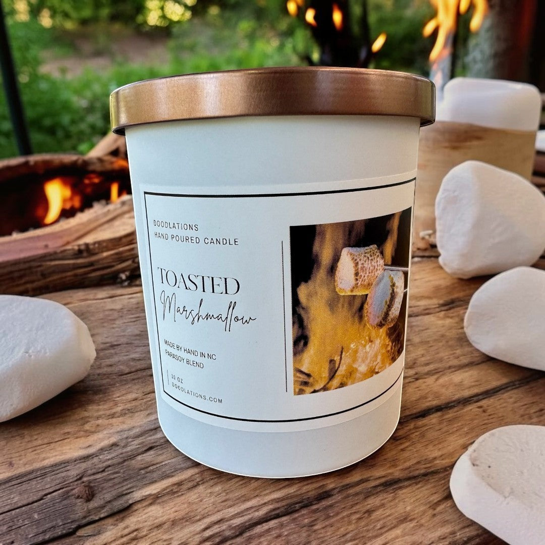 Doodlations Toasted Marshmallow hand-poured candle with a copper lid, set on a wooden table near a campfire.