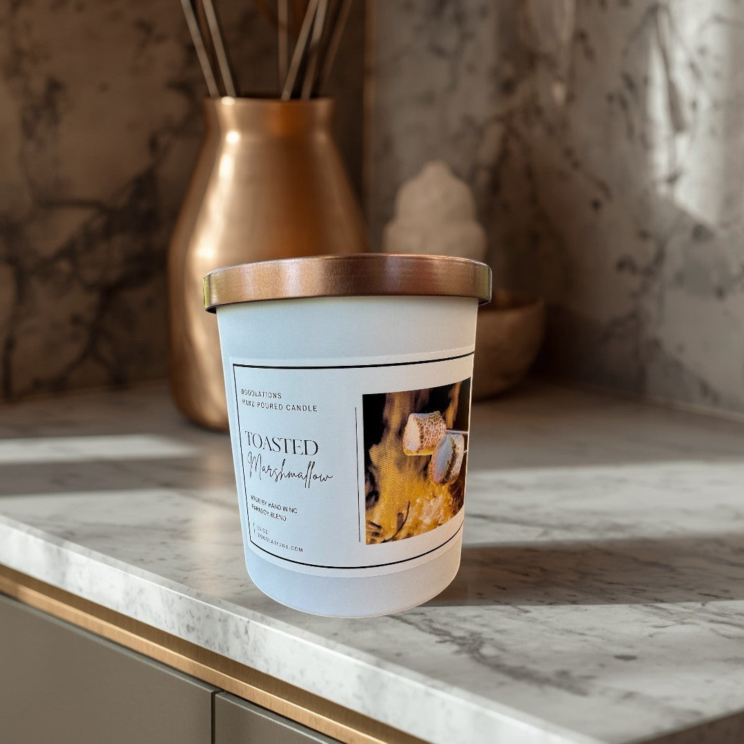 Doodlations Toasted Marshmallow hand-poured candle with copper lid, set by a campfire with marshmallows.
