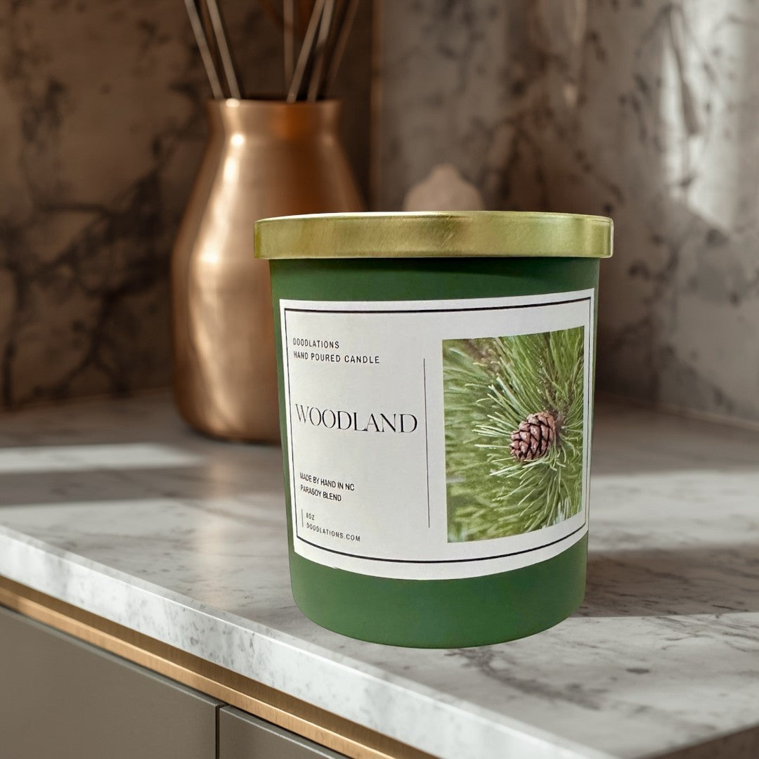 Doodlations Woodland hand-poured para-soy blend candle in a green jar with a gold lid, featuring a pine cone image.