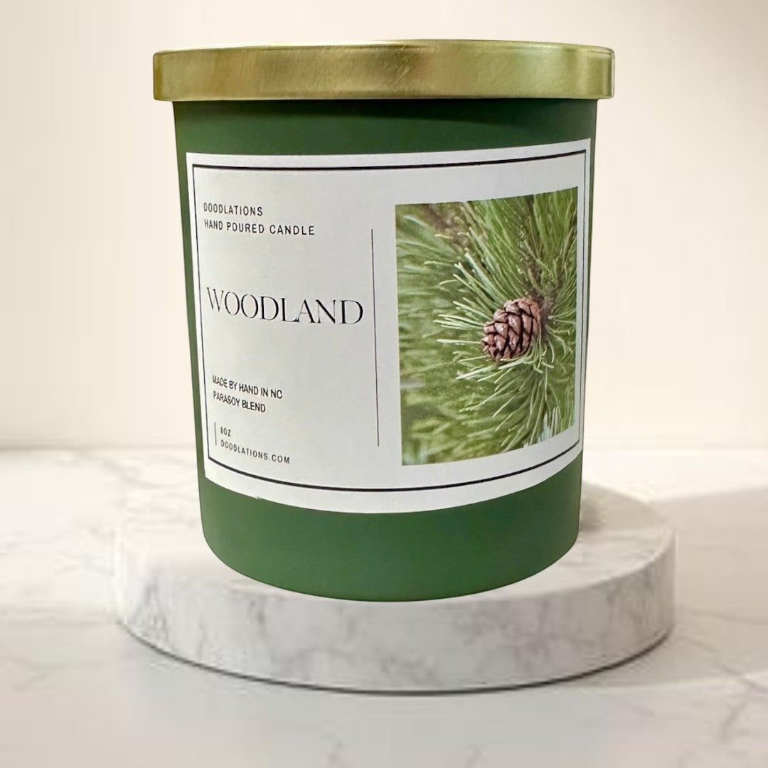 Doodlations Woodland hand-poured para-soy blend candle in a green jar with a gold lid, featuring a pine cone image.
